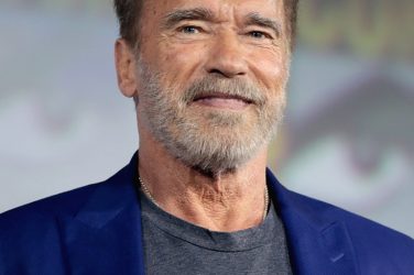 Picture of Arnold Schwarzenegger at the age of about 70 years old with scraggly gray beard, aged skin, receding hairlkne, slight smile, direct eye contact, gray t-shirt, blue blazer