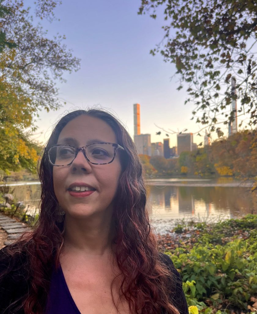 Headshot of woman in urban park with tall buildings behind her
